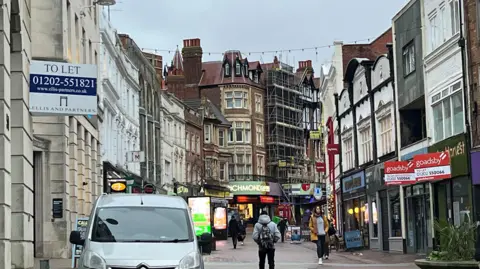 BBC A high street with people walking. To let and signs above some of the empty shops on the high street.