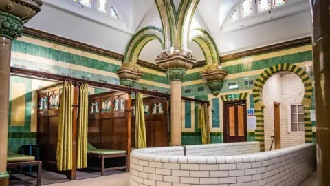 BBC Carlisle's Turkish Baths, pictured before the closure. The walls are tiled in green and white. There are three small, curtained cubicles to the left with wooden benches in each one.