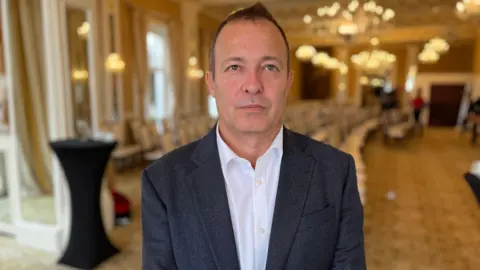 A man with short hair looking at the camera wearing a white shirt and a blue suit. 