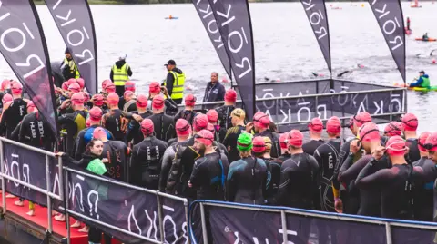 Roger Trice/Ironman Swimmers line up in matching hats and wetsuits in front of a large lake. There are marshalls wearing hi-vis jackets.