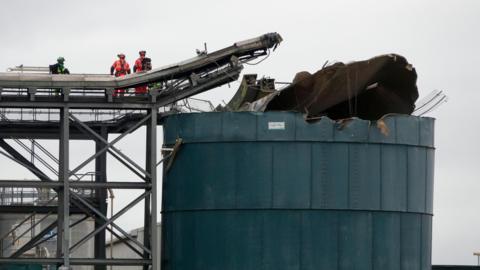 Image of the explosion at Avonmouth water recycling centre. Emergency service workers wearing high vis can be seen next to a damaged structure.