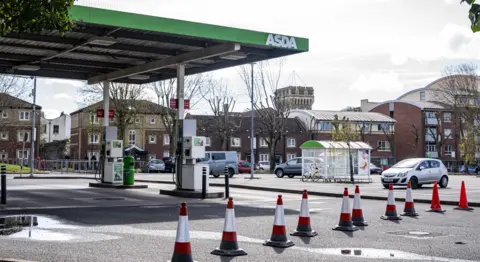 Ben Birchall/PA Wire The entrance to an Asda forecourt is blocked off by a row of orange traffic cones. The forecourt is empty but a few cars are parked in the bays behind the forecourt including a silver Corsa. There are some small puddles on the ground which are reflecting the sunlight.
