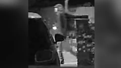 Sussex Police Grainy nighttime CCTV images of a man in a cap walking in front of a brick building with some bins in front of it. 
