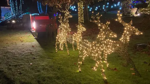 Six illuminated reindeer sculptures are styled to appear to be towing a red sleigh. They are in a garden with hedges and trees also illuminated.