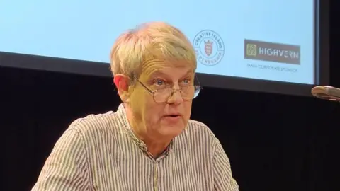 A man, in a white and purple striped top. He is looking forwards and his mouth is slightly open. He is wearing glasses. Behind him is a projector screen and black curtain. 