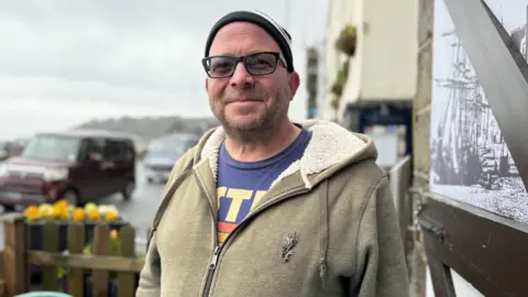 A middle aged man with glasses and a black and white beany style hat on. He is wearing a stone brown cardigan with a wooly style inside hood. He has a purple t-shirt on with yellow writting.