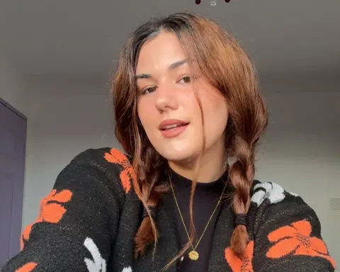 Summer-Rose Starenczak A woman with her hair in two plaits looks at the camera. She is wearing a black cardigan with orange and white flowers and is stood indoors