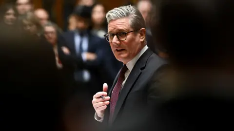 PM Sir Keir Starmer speaks during Prime Minister's Questions. The camera is positioned between MPs, who are blurred out in the foreground, while Starmer appears clearly between them in the background. 