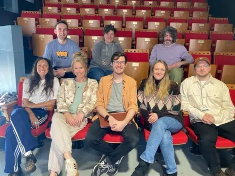 Eight people smile as they sit in two rows at the front of an intimate theatre auditorium. They are wearing casual clothes. The theatre seats are made of brown wood with red velvet seats and backs. Six rows of seats can be seen.