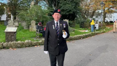 BBC/George Carden Tony Avery wearing a dark blazer, service medals and beret, standing to attention in front of Shoreham's war memorial.