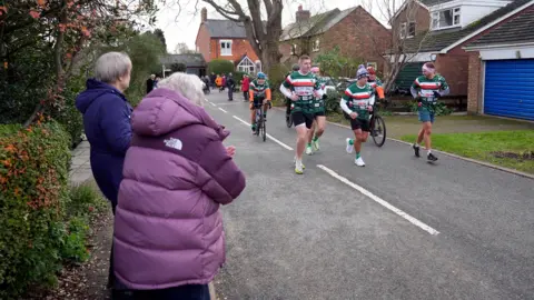 PA A man in a blue jacket and a woman in a purple jacket clap as a pack of runners wearing green, red, white striped rugby shirts run down a road of a housing estate.