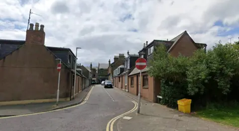 A terraced residential street with No Entry signs and a grassy area with trees to the right of the photo.