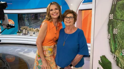 Savannah Guthrie, in a bright orange sleeveless top and patterened orange hued skirt, smiles beside her mother Nancy, in dark-rimmed glasses and a deep blue T-shirt, on a TV news set