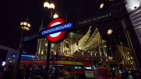 Getty Images The entrance to Piccadilly Circus underground station is pictured at night, in front of a large illuminated angel strung across Regent Street. Two red London buses pass through the shot. 