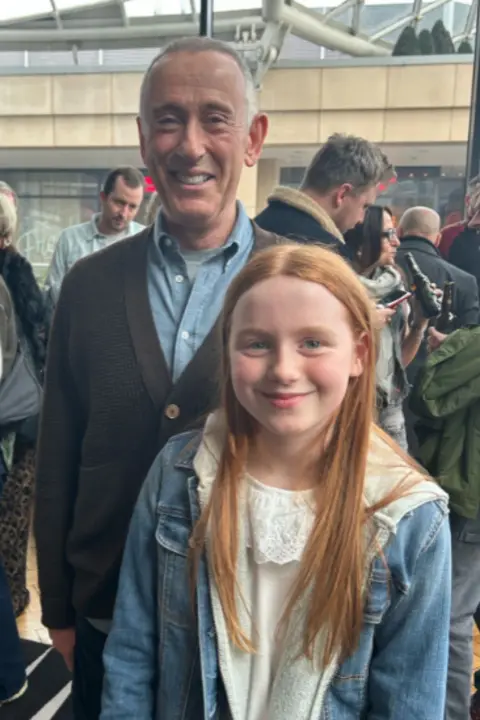 Sarah Armitage Nicholas Hytner and Anna Cook look at the camera and smile, with a group of people in the background.