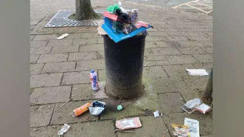 Tim Manton There is a tall black bin which is overflowing with litter. On the floor is rubbish including a Yazoo bottle, an orange boost bottle, a gregs wrapper and a haribo goldbears wrapper. On top of the bin, there is rubbish piled on top of a big Dominos box.