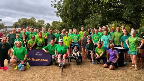 Carrol Weston/BBC At least 45 people, a mix of men and women, lined up, mostly wearing green T-shirts. One man in the middle is pushing a buggy with a child in it and a woman is using a sport