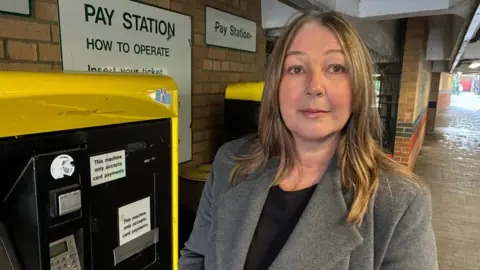 Woman in grey jacket with blonde hair stood in a car park by a parking metre. 
