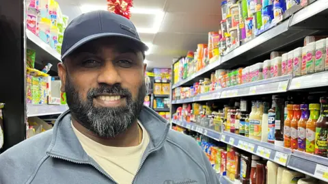 Raj Thurairaja is standing in his convenience store with products packed on the shelves behind him. He is smiling and wearing a black cap and a grey jacket and cream T-shirt. 