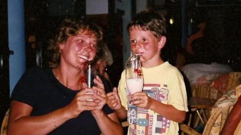 A woman smiling at a young boy sipping milk from a glass through a straw