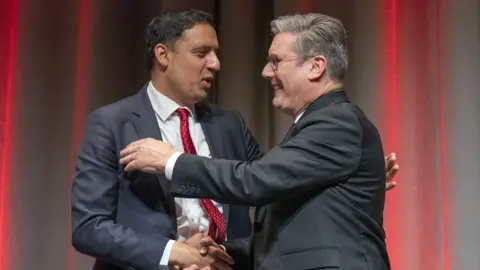 PA Media Anas Sarwar, with dark hair and wearing a navy suit, embraces Sir Keir Starmer, with grey hair and a black suit. They are on stage, with curtains illuminated by red lights in the background. 