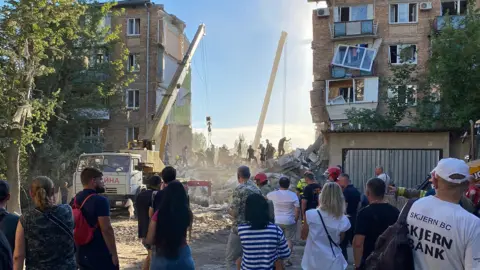 Leila Osman A building has been destroyed and cranes and workers are trying to move the rubble. A crowd has gathered to watch.