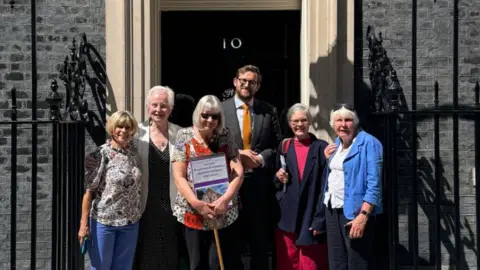 Freddie van Mierlo in a grey suit, blue shirt and orange tie, along with five women all with grey hair.