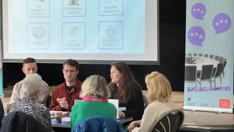 Two men and four women sit around a table at Seaton Delaval arts centre. They are talking to each other and all have serious expressions. Behind them a slide is projected on a screen on a stage with guidelines on how to conduct discussions.