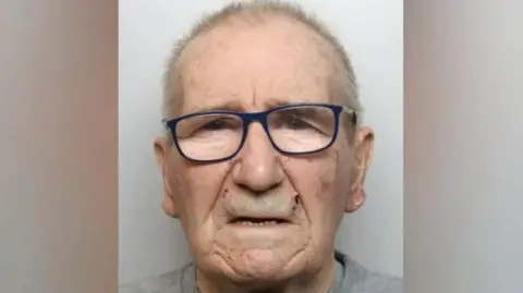 Peter Matthews, an elderly man with short hair and glasses, is wearing a grey jumper. He is posing for a police mugshot.