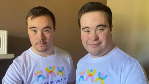 Two young men are smiling as they stand side by side in a house, both wearing white T-shirts with the pink, yellow and blue Down's Syndrome Manchester logo on.