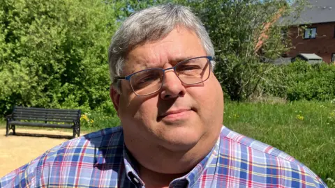A man with short grey hair and glasses, wearing a blue, red and white check shirt, standing in a garden on a sunny day.