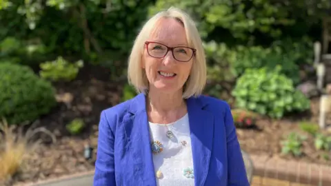 The image shows Christine Chapman standing in her garden. She is looking at the camera and smiling. There are green shrubs behind her, and she is wearing a white top and a blue jacket.