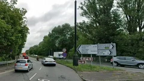 Google Dual carriageway with traffic on both sides of the road and several signs on the central island