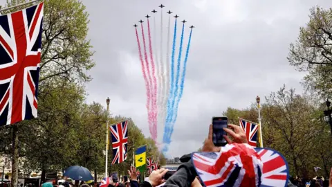 Reuters The Red Arrows make a flypast up The Mall, during the coronation ceremony of King Charles and Queen Camilla, in 2023. The distinctive smoke trails of the Red Arrows, in red, white, and blue colours, can be seen. In The Mall there are flags hanging from lampposts and crowds are taking pictures of the flying display.