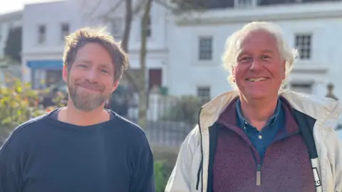 Two men staring directly at the camera. On the left, the man is wearing a blue Guernsey jumper. He has a beard and short brown hair. On the right, the man is wearing a maroon, quarter-zip jumper over a blue shirt. He is also wearing a white raincoat, which is unzipped. He has short white hair.