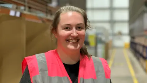 A woman with tied back brown hair and a pink reflective jacket in a blurred room