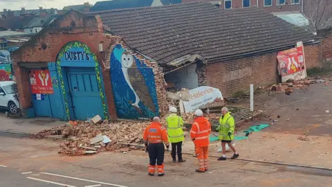 Alan Bolster Old building with section of brick wall demolished. There is a pile of bricks in front of the wall and four people - wearing construction-type clothing are standing next to the building. A picture of an owl is painted on the building front wall and the name Dusty's is written above the front doors.