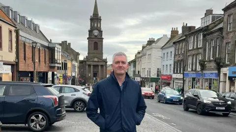 David Smith David Smith is a man in his 40s, clean-shaven with greying hair wearing a blue jacket with his hands in his pockets. He is standing in Berwick's high street with cars parked on one side and a row of shops going down the hill to a clock tower at the bottom.