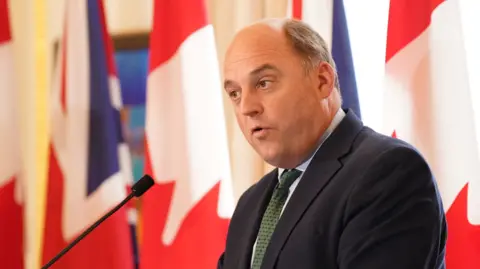 Image shows former Defence secretary Ben Wallace, who is partially bald and is wearing a suit, standing on a podium in front of a number of unfurled Union Jacks.
