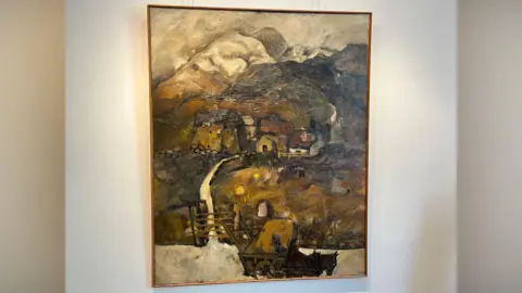 Pathway to a Farm framed in a thin wooden frame, hanging from a wall. It shows a rural scene, with a horse and carriage in the foreground in front of a farm gate opening to a path leading to a cluster of buildings. In the background are snow-capped mountains. 