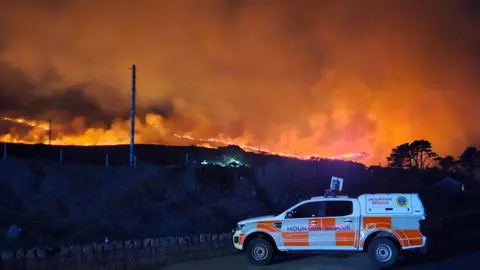 Mourne Mountain Rescue Team A Mourne Mountain Rescue Team 4x4 vehicle parked in the foreground of a large wildfire at night. The liveried vehicle is white and orange with a large "mountain rescue" sign in blue lettering. A line of fire stretches across a mountain in the background, illuminating large plumes of smoke which appear orange against the night sky. 