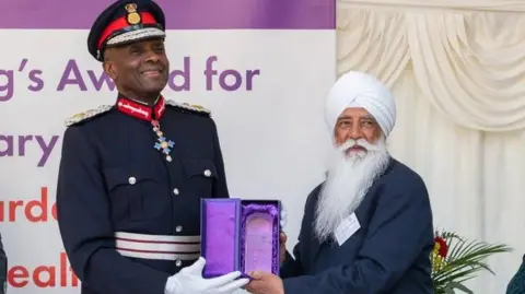 Nishkam Healthcare Trust A man in a black military jacket with gold epaulets and red and white striped band around the middle with cross hanging from his neck and red and black hat presents an award in a purple box to a another man on his right. The second man is smaller and wears a white turban and has a moustache and long white beard.