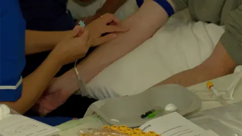 A patient is about to receive a blood test. A nurse is holding a needing in one hand and has placed her finger of the vein on a man's arms. The man is sitting down and is resting his arms on a pillow in a hospital setting. 