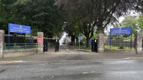 Two signs saying 'Whitchurch High School' are on either side of a driveway with gates. A school building can be seen further down the drive.