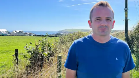 Gareth Williams stands in a blue T-shirt next to a field with houses and the sea in the background