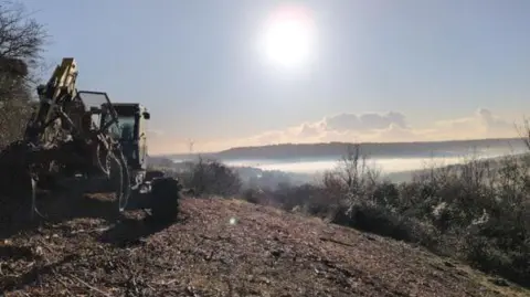 National Highways Image of a digger in a brown field at the top of a hill. In the background can be seen a view of hills and countryside and a valley shrouded in mist.