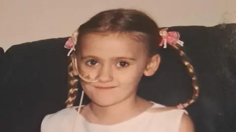 Demie Risby A young girl with her blonde hair in braided pig tails. She has a white tube up her nose.