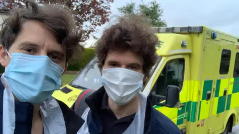 Contributed Ryan and Dean Appleton take a selfie style image during the Covid pandemic. They both are wearing face masks. An ambulance can be seen behind them.