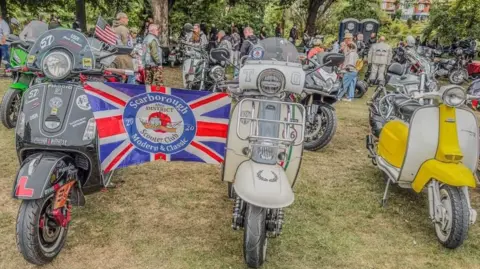Three scooters in a row with a Union flag with the words Scarborough Modern and Classic on it. There are other motorcycles in the background with people gathered around.