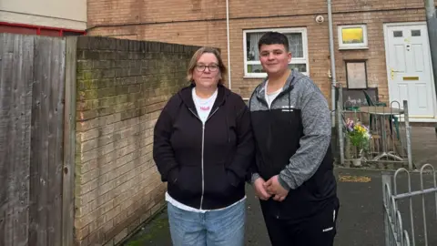 A woman and a teenager stand side by side next to a wall in front of a block of flats behind them. She is wearing a black hoodie, a white t-shirt and jeans, and has her hands in her pockets. He is wearing a black and grey tracksuit.